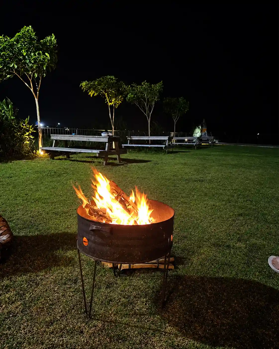 Fire burning in an outdoor fire pit on the lawn at night, with picnic benches behind.